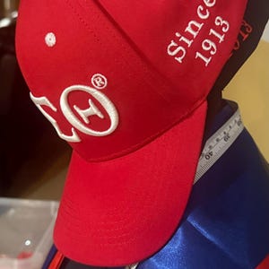 Op de afbeelding: Rode baseballcap met witte geborduurde details, waaronder de tekst "Since 1913" en een logo. De cap heeft een gebogen klep en een witte knoop bovenop. Een meetlint is zichtbaar.