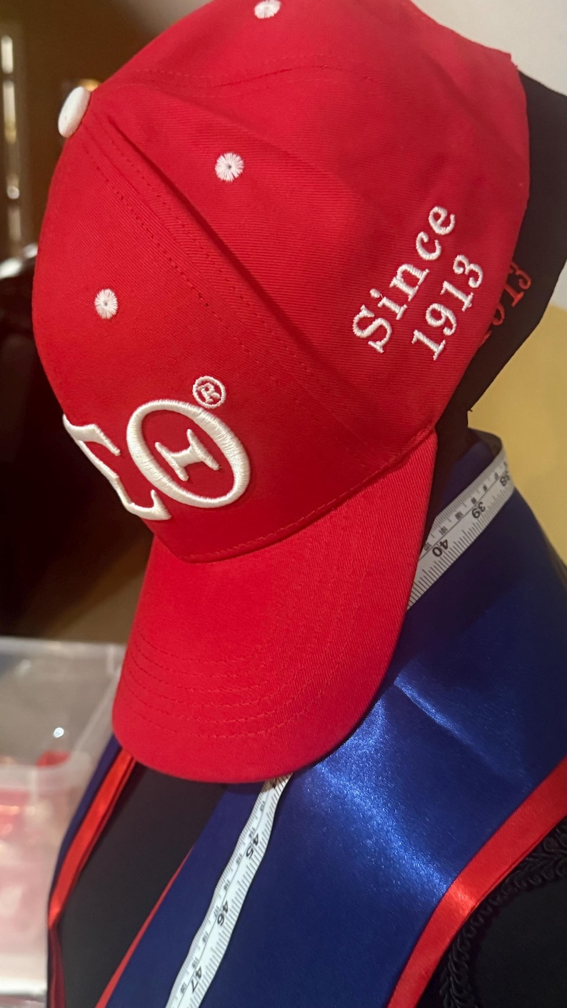 Op de afbeelding: Rode baseballcap met witte geborduurde details, waaronder de tekst "Since 1913" en een logo. De cap heeft een gebogen klep en een witte knoop bovenop. Een meetlint is zichtbaar.