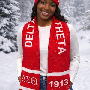 Puede incluir: Un gorro rojo de punto con una banda blanca y una bufanda a juego. El gorro tiene un pomp&oacute;n y las letras griegas Delta Sigma Theta. La bufanda tiene las mismas letras y el a&ntilde;o 1913.