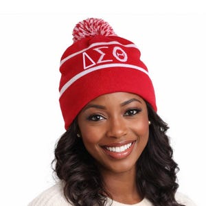 Puede incluir: Gorro de punto rojo con rayas blancas y un pomp&oacute;n blanco y rojo. El gorro presenta letras griegas blancas. Un accesorio de invierno c&aacute;lido y elegante.