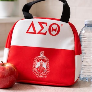 Op de afbeelding: Een rode en witte lunchtas met een zwart handvat. De tas toont de Griekse letters &Delta;&Sigma;&Theta; in rood op een witte achtergrond. Daaronder staat een wapen met Griekse letters en een fakkel op het rode deel van de tas. Een appel en een waterfles staan naast de tas.