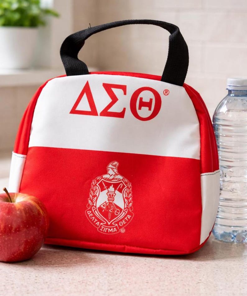 Op de afbeelding: Een rode en witte lunchtas met een zwart handvat. De tas toont de Griekse letters &Delta;&Sigma;&Theta; in rood op een witte achtergrond. Daaronder staat een wapen met Griekse letters en een fakkel op het rode deel van de tas. Een appel en een waterfles staan naast de tas.