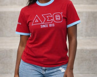 Puede incluir: Una camiseta roja con ribetes azul claro en el cuello y las mangas. La camiseta presenta el texto blanco "ΔΣΘ SINCE 1913". La modelo lleva vaqueros azules rotos.