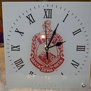 May include: Square white clock with black Roman numerals and black hands. The clock face features a red and white emblem with Greek letters. A small, clear gem is set in the lower right corner.