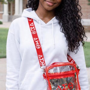 Op de afbeelding: Een doorzichtige rode crossbodytas met een rode band met witte Griekse letters. De tas heeft een rode rits en een rode band met witte Griekse letters. De tas wordt gedragen over een witte hoodie.