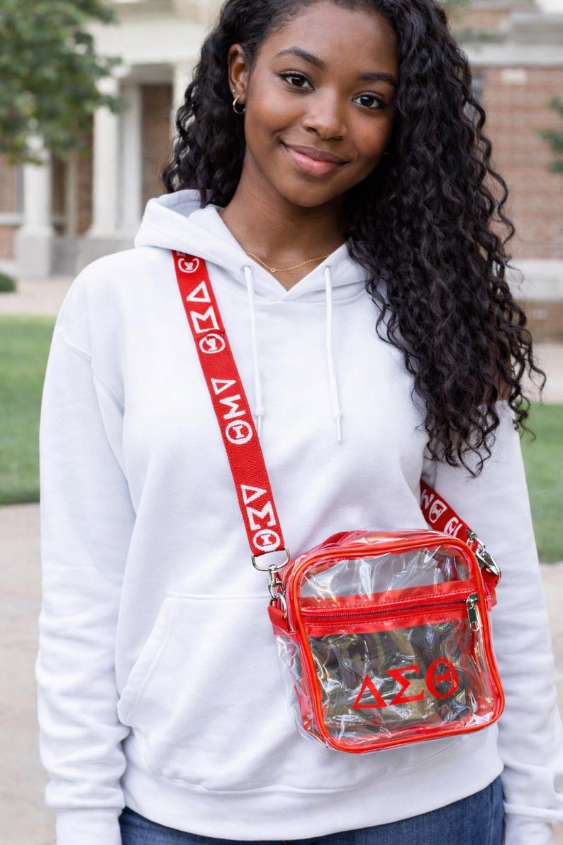 Op de afbeelding: Een doorzichtige rode crossbodytas met een rode band met witte Griekse letters. De tas heeft een rode rits en een rode band met witte Griekse letters. De tas wordt gedragen over een witte hoodie.