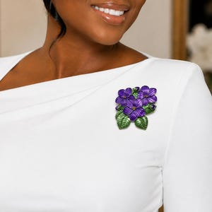 May include: A floral brooch featuring three purple flowers with yellow centers and green leaves. The brooch is attached to a white garment. The flowers have a textured appearance.