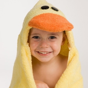 May include: A yellow hooded towel with a duck face design. The hood features black eyes and an orange beak. The towel is wrapped around a child, creating a playful and fun look. The child is smiling.