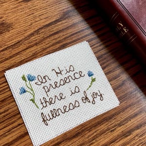 Puede incluir: Un pequeño punto de cruz blanco con las palabras "In His presence there is fullness of joy" en hilo marrón. Acentos florales azules adornan los bordes. Una Biblia marrón encuadernada en cuero descansa en el fondo sobre una superficie de madera.