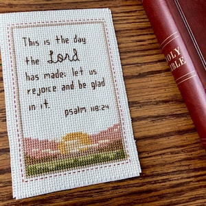 Könnte beinhalten: Ein kreuzgesticktes Lesezeichen mit dem Text „Dies ist der Tag, den der HERR gemacht hat; lasst uns fröhlich sein und uns freuen. Psalm 118,24.“ Das Lesezeichen zeigt eine Landschaft mit Sonnenaufgang. Eine rotlederne Bibel ist teilweise sichtbar.