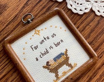 Może przedstawiać: Oprawiony haft krzyżykowy z napisem "For unto us a child is born" i przedstawieniem żłóbka. Rama jest drewniana. Tło zawiera szpulki nici i białą koronkową serwetkę.
