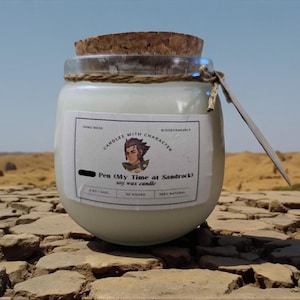 May include: A soy wax candle in a clear glass jar with a cork lid. The label reads "Candles with Character" and features an illustration. The candle is labeled "Pen (My Time at Sandrock)" and is 5.62 oz. The background is a desert landscape.