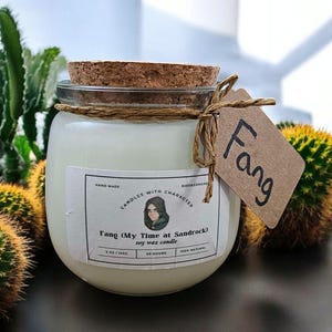May include: A white soy wax candle in a clear glass jar with a cork lid. The jar has a label that says "Fang (My Time at Sandrock) soy wax candle" and "Hand Made Candles with Character". The candle is tied with twine and has a brown paper tag that says "Fang".