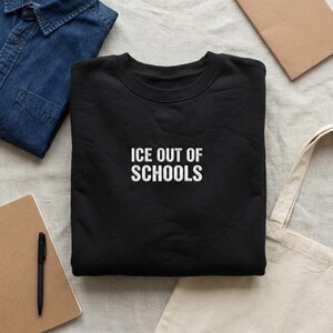 May include: Black crew neck sweatshirt with the white text "ICE OUT OF SCHOOLS" centered on the front. The sweatshirt is folded on a light-colored surface, with a denim shirt, notebook, pen, and tote bag in the background.