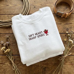 May include: White sweatshirt with the embroidered text "SOFT HEART, SHARP SPURS" and a cartoon heart wearing a cowboy hat. The sweatshirt is folded on a wooden surface, with a rope and belt in the background.