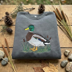 May include: A grey sweatshirt featuring an embroidered mallard duck. The duck has green, brown, white, and blue details, set in a grassy area with bulrushes. The sweatshirt is displayed on a wooden surface with natural decorations.