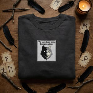 May include: Dark grey sweatshirt with a patch showing a black and grey wolf design and the text "We were born from the same night." The image is surrounded by feathers, runes, twigs, and a lit candle.