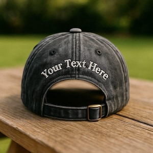 May include: A black baseball cap with the text "Your Text Here" embroidered in white. The cap has a curved brim, a metal buckle closure, and a small button on top. The cap is sitting on a wooden surface.