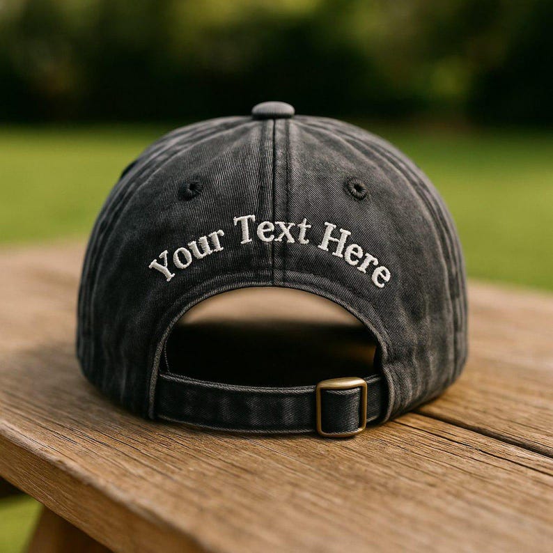 May include: A black baseball cap with the text "Your Text Here" embroidered in white. The cap has a curved brim, a metal buckle closure, and a small button on top. The cap is sitting on a wooden surface.