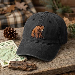 May include: A charcoal grey baseball cap featuring an embroidered patch of a brown bear sitting down. The cap has a structured crown, a curved brim, and stitched eyelets on the top panels. The bear design is rendered in shades of brown and tan.