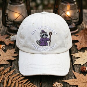 May include: A cream-coloured baseball cap with the embroidered text "OFF TO COMMIT SHENANIGANS". The cap features an embroidered raccoon in a purple wizard robe and hat, holding a staff. The hat is distressed and has a curved brim.
