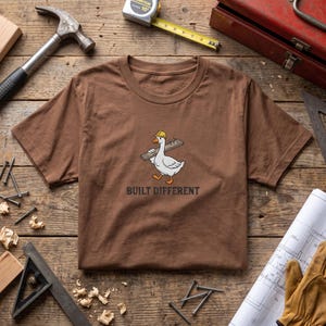 May include: A brown t-shirt featuring a graphic of a white goose wearing a yellow construction hard hat and carrying a wooden board with nails. The text "BUILT DIFFERENT" is printed in black capital letters below the goose illustration.