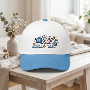 May include: White baseball cap with a light blue brim and top button. The cap features an embroidered design of three cartoon creatures, clouds, stars, and a crescent moon. The creatures are in blue and pink.