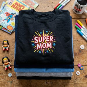 May include: A stack of four sweatshirts in black, navy, light blue, and gray. The top black sweatshirt features a colorful "SUPER MOM" graphic. Comic books, Wonder Woman figurines, and a "Best Mom Ever" mug are also visible.