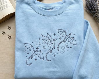 May include: A light blue sweatshirt featuring a black embroidered design of three dragons with outstretched wings, stars, and crescent moons. The sweatshirt is made of a soft material and has a crew neck.