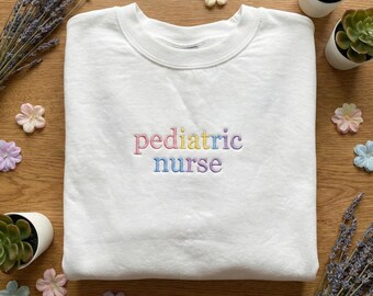 May include: A white crewneck sweatshirt with the embroidered text "pediatric nurse" in pastel colors. The sweatshirt is surrounded by decorative elements, including small flower-shaped decorations, lavender sprigs, and small potted succulents.