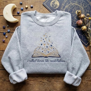 May include: A grey sweatshirt with a crew neck and rolled cuffs. It has an embroidered open book design with blue and gold stars and the text "i collect stories like constellations".