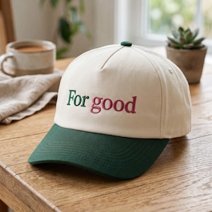 May include: A cream-coloured baseball cap with a dark green brim and a dark green button on top. The front of the cap features the embroidered text "For good" with "For" in dark green and "good" in a deep pinkish-red colour.
