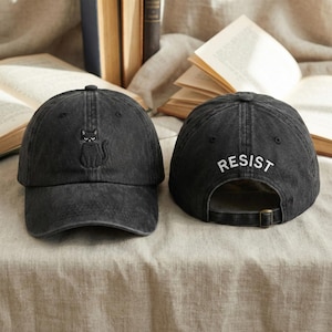 May include: Two black baseball caps. One has a black embroidered cat, the other has the word "RESIST" embroidered in white. Both caps have a curved peak and an adjustable strap. The caps are displayed on a neutral-toned surface.