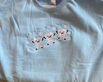 May include: A light blue crewneck sweatshirt featuring an embroidered design of three smiling white teeth characters skiing on a white line, surrounded by small white embroidered snowflakes across the centre of the chest.