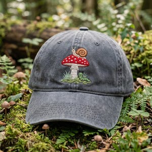 Peut inclure: Une casquette de baseball gris anthracite ornée d'une broderie représentant un champignon rouge à pois blancs surmonté d'un escargot brun, avec une petite chenille verte rampant sur le sol à la base de la tige du champignon.