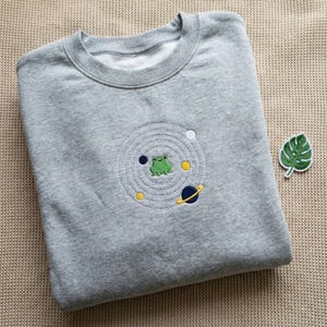 May include: A grey sweatshirt with an embroidered design of a green frog within a planetary system. The design includes white, yellow, and blue stitching. A green leaf patch is also visible.