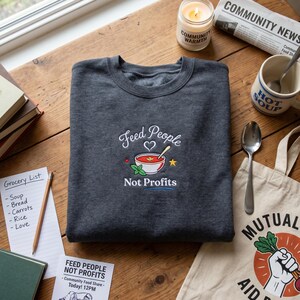 May include: A dark gray sweatshirt with the embroidered text "Feed People Not Profits" above a bowl of soup. The image includes a grocery list, a candle, a mug that says "Hot Soup", and a tote bag.