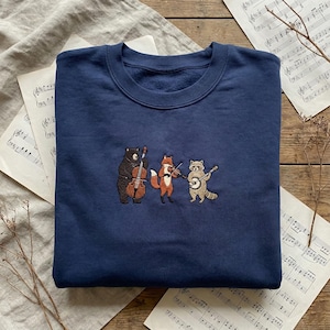 May include: Navy blue sweatshirt featuring an embroidered design of a bear playing a cello, a fox playing a violin, and a raccoon playing a guitar. The musical animals are on a navy blue background. Sheet music and dried plants are in the background.