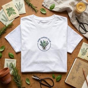 Peut inclure: Un t-shirt blanc à col rond avec un motif circulaire "Herb Garden Club" représentant un bouquet d'herbes. Le t-shirt est posé sur une surface en bois avec des sachets de graines d'herbes, un carnet et des ciseaux.
