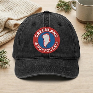 May include: A black denim baseball cap with a circular patch featuring the text "GREENLAND IS NOT FOR SALE" around a map of Greenland. The patch has a red border, blue center, and white map outline.