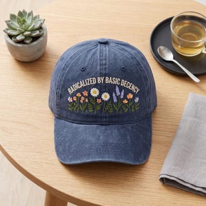 May include: A navy blue baseball cap featuring an embroidered design of colourful wildflowers and the text "RADICALIZED BY BASIC DECENCY" in a light cream font. The cap has a distressed, washed-out texture and a curved brim with stitching.