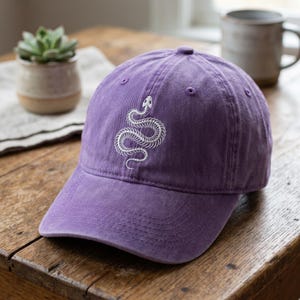 Peut inclure: Une casquette de baseball violette avec un motif brodé blanc représentant un squelette de serpent sur le panneau avant. La casquette présente une visière incurvée, un bouton sur le dessus et des œillets cousus pour la ventilation.