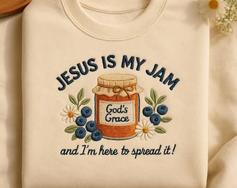 May include: Cream-colored sweatshirt with embroidered text and graphics. The design features the phrase "JESUS IS MY JAM" above a jar of jam, blueberries, and daisies. Below the jar is the phrase "and I'm here to spread it!"