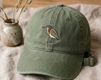 Puede incluir: Una gorra de béisbol de color verde oliva con un gráfico bordado de una garza verde en el panel frontal. La gorra tiene una visera curva, ojales cosidos y una correa de tela ajustable con un cierre de hebilla de metal en la parte posterior.