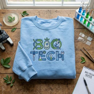 May include: A light blue sweatshirt with the words "BIO TECH" in a science-themed design. The design includes a DNA strand, a microscope, and other lab equipment. The sweatshirt is on a wooden surface with lab equipment.