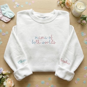 Puede incluir: Sudadera blanca con bordado "mama of both worlds" en azul y rosa. Las mangas llevan bordados los nombres "Ethan" y "Sophie". Confeti y flores en tonos pastel decoran la imagen.