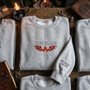 May include: Gray sweatshirt with the text "IRON FLAME" above a red and yellow flame design. The sleeve has the text "We choose our fate". The sweatshirts are displayed on a wooden surface.
