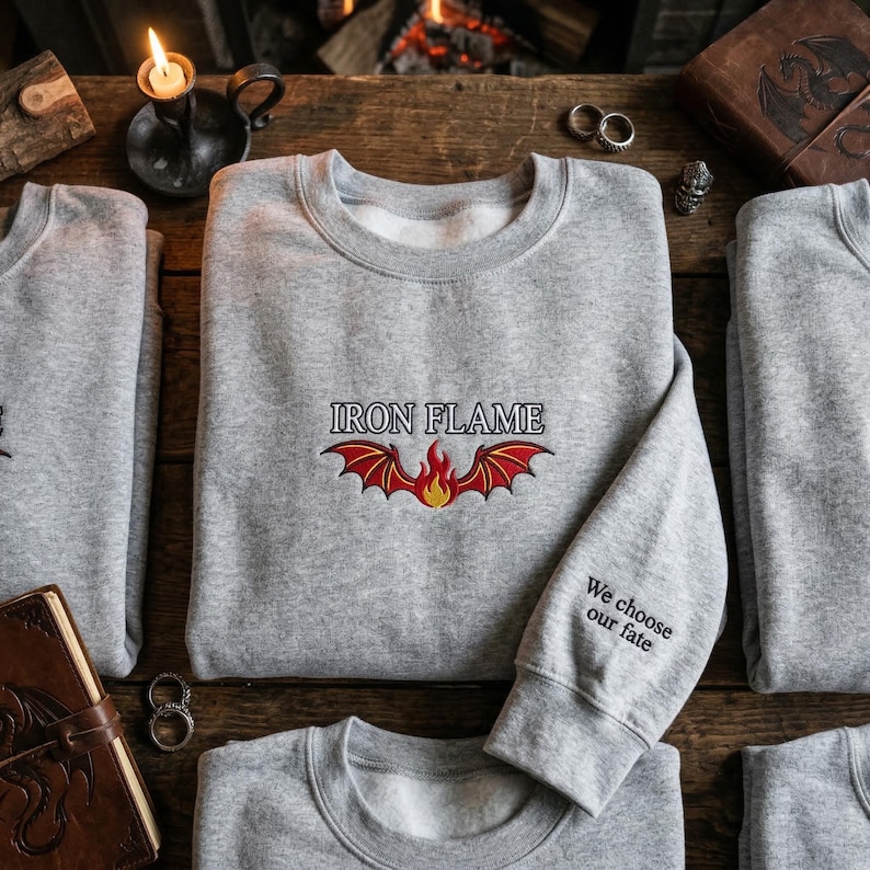 May include: Gray sweatshirt with the text "IRON FLAME" above a red and yellow flame design. The sleeve has the text "We choose our fate". The sweatshirts are displayed on a wooden surface.