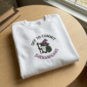 May include: White crew neck sweatshirt with an embroidered design. The design features a green frog wearing a purple wizard hat and holding a staff, with the text "OFF TO COMMIT SHENANIGANS" in green and purple.