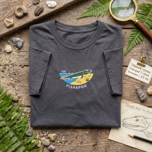 May include: A dark gray t-shirt featuring an embroidered graphic of a green Tiktaalik fish swimming in blue water above a yellow sandy shore. The text FISHAPOD is embroidered in white below the graphic. The shirt has a crew neckline.